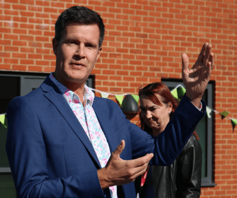 Richard Kennett, CEO of The Loddon School, speaking outdoors in front of the new school building decorated with green bunting. Wearing a blue suit and a vibrant patterned shirt, he gestures expressively with his hands while addressing an audience. A smiling staff member in a black leather jacket stands just behind him.