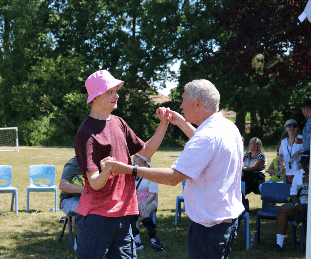 Two individuals joyfully dancing together outdoors on a sunny, grassy field at The Loddon School. A young man wearing a bright pink bucket hat and a maroon t-shirt is holding hands and dancing with an older man with grey hair in a light-striped shirt. In the background, several other people are relaxing on light blue plastic chairs, enjoying the outdoor gathering near a line of trees