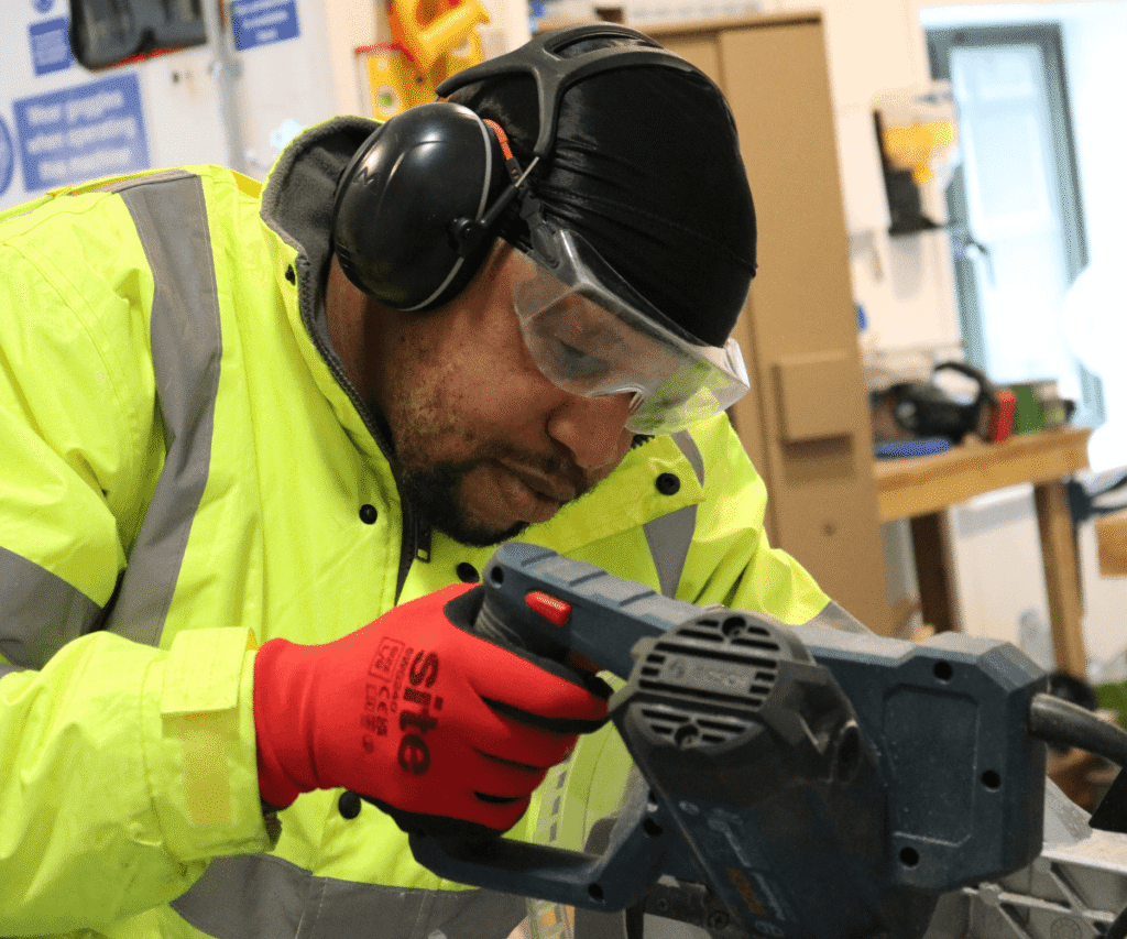 A staff member at The Loddon School carefully operating a saw in a workshop. He is focused on his work and wearing full personal protective equipment, including a bright yellow high-visibility jacket, red safety gloves, clear safety goggles, and black ear defenders over a black head covering.