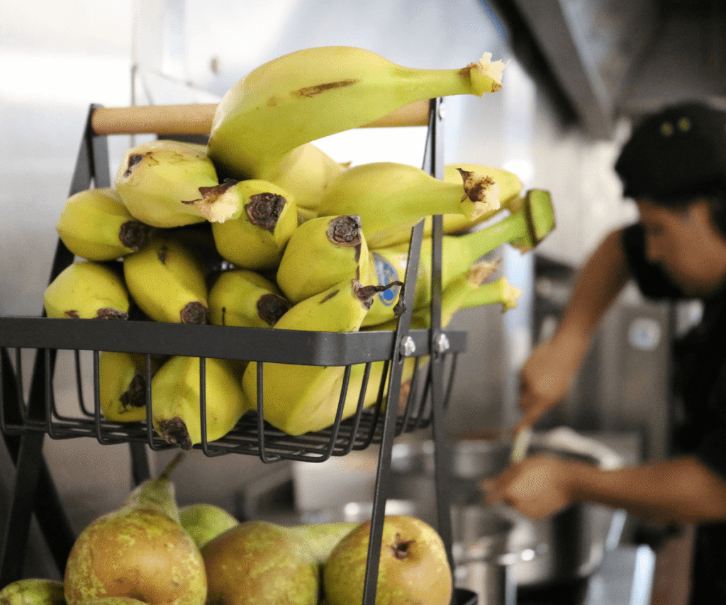 A close-up of a black wire, two-tiered fruit basket in one of The Loddon School's kitchens. The top tier is filled with a bunch of slightly green bananas, and the bottom tier holds several pears. In the blurred background, a staff member in a black uniform is working, stirring food in a large metal pot.