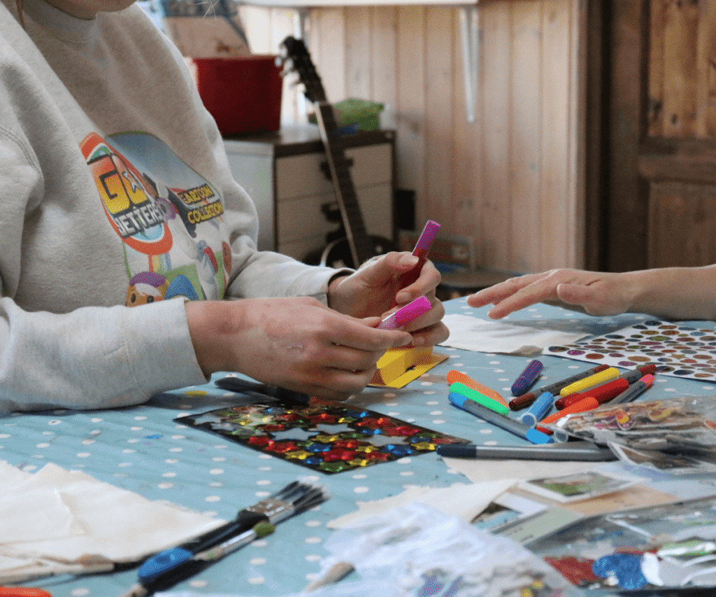 Two people sitting at a table with a light blue polka-dot tablecloth at The Loddon School, engaged in an arts and crafts activity. One person, wearing a grey cartoon sweatshirt, holds a pink marker. The table is scattered with colourful markers, shiny star stickers, a star stencil, and paintbrushes.