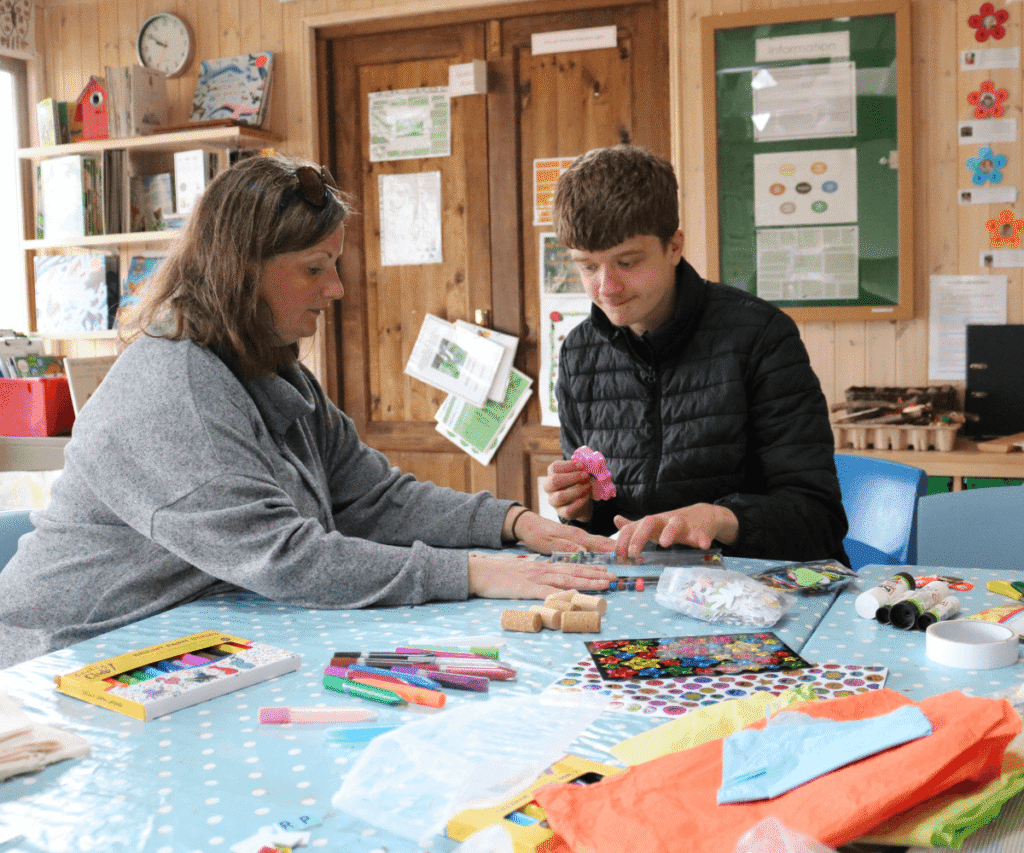 A staff member and a student sitting together at an indoor craft table at The Loddon School. The student, wearing a black puffer jacket, holds a pink moulded shape while the staff member, in a grey top, helps organise the materials. The blue polka-dot table is covered with craft supplies including markers, paint sticks, glue, corks, and colourful tissue paper.