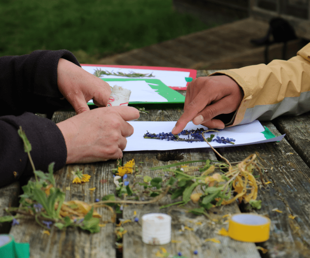 A close-up of two pairs of hands working on a nature craft project at an outdoor wooden picnic table at The Loddon School. One person is applying glue while the other points to a piece of paper decorated with small pressed blue flowers. Scattered across the table are freshly picked wildflowers, leaves, and rolls of yellow and green tape.