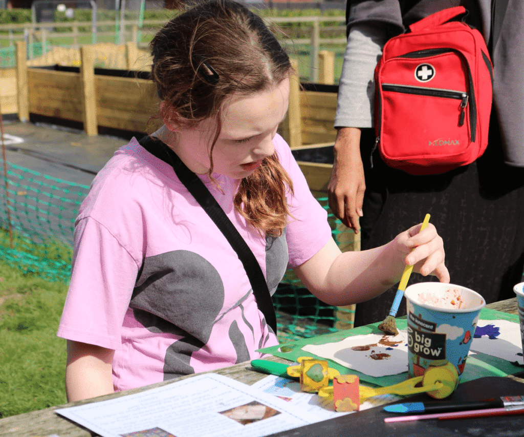 A child at The Loddon School sits at an outdoor wooden table, focused on a painting activity in the sunshine. Wearing a pink t-shirt, she uses a paintbrush to dab brown paint onto paper. The table is scattered with craft supplies, including yellow sponge rollers and a printed activity sheet. A staff member stands behind her, carrying a bright red first aid backpack. Wooden raised garden beds and green fencing are visible in the background.