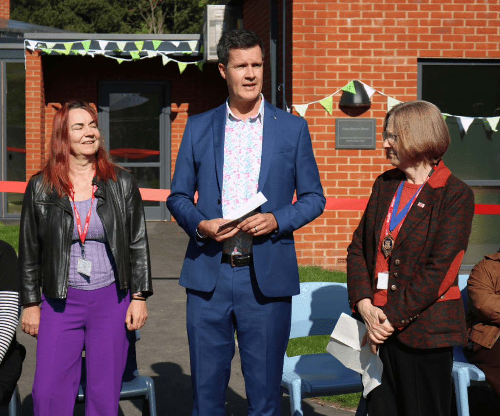 Three individuals standing outdoors during a building opening ceremony at The Loddon School. In the centre is Richard Kennett, CEO, wearing a blue suit and patterned shirt, holding notes and speaking. To his left stands a smiling woman with red hair in a black leather jacket and vibrant purple trousers. To his right is another woman in a patterned red and black jacket, wearing a large silver civic medal on a blue ribbon. A red ribbon stretches across the space behind them in front of a modern red brick building decorated with green and white bunting.