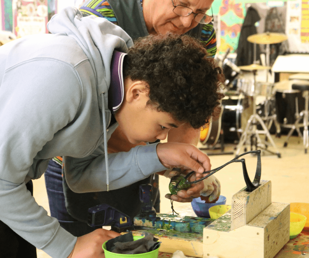 A student wearing a light blue hoodie leans in closely to examine a green painted object clamped to a table in a room at The Loddon School. A staff member wearing glasses and a brightly striped shirt stands behind him, closely observing his work. A drum kit is slightly blurred in the background.