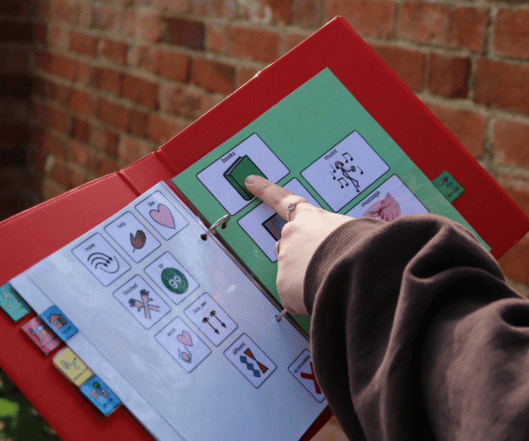 A close-up of a person's hand pointing to a 'books' symbol inside an open red visual communication folder at The Loddon School. The page displays various other picture communication symbols, including 'help', 'more', 'music', and 'massage', designed to aid in expression and choice-making.
