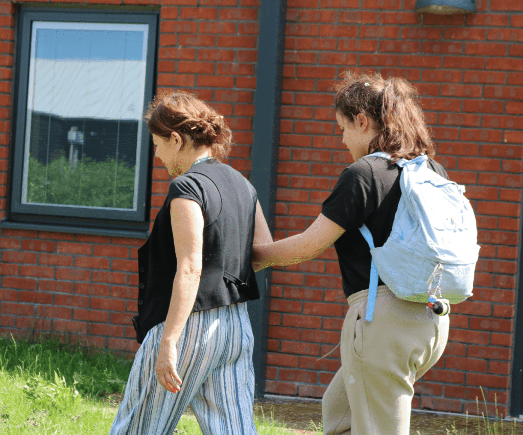 A staff member and a child at The Loddon School walking together on the grass alongside a red brick building. The child, wearing a black t-shirt, beige joggers, and carrying a light blue backpack, is gently holding the arm of the staff member, who is wearing a black top and blue-and-white striped trousers.