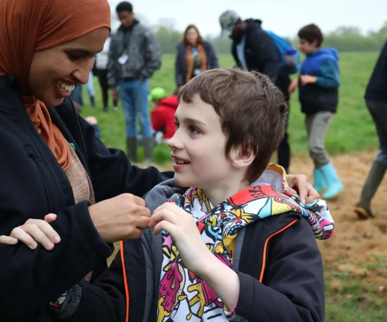 A woman wearing a brown hijab and a black jacket smiles warmly as she holds the arms of a young boy looking up at her. The boy is wearing a brightly coloured hoodie with cartoon patterns. They are standing in a grassy field, and a group of people wearing casual outdoor clothing and rain boots can be seen blurred in the background.