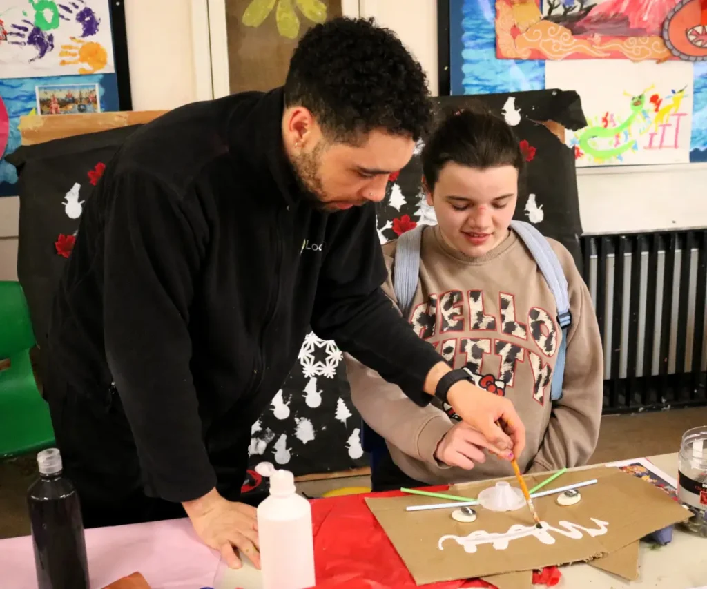 A staff member at The Loddon School stands beside a young person, gently guiding her hand as she uses a paintbrush to apply white paint to a cardboard craft project. The young person is smiling and wearing a Hello Kitty sweatshirt with a light blue backpack. They are working at a table with various art supplies, and colourful children's artwork is displayed on the walls in the background.