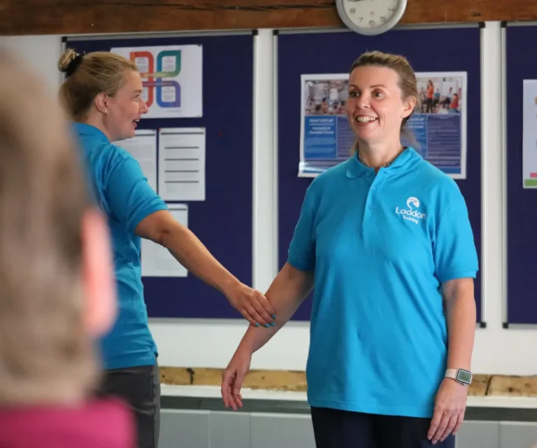 Two female instructors in blue Loddon Training polo shirts demonstrate a positive behaviour support technique. One smiles towards the audience while the other stands beside her, gently supporting her forearm in a training space with exposed wooden beams.