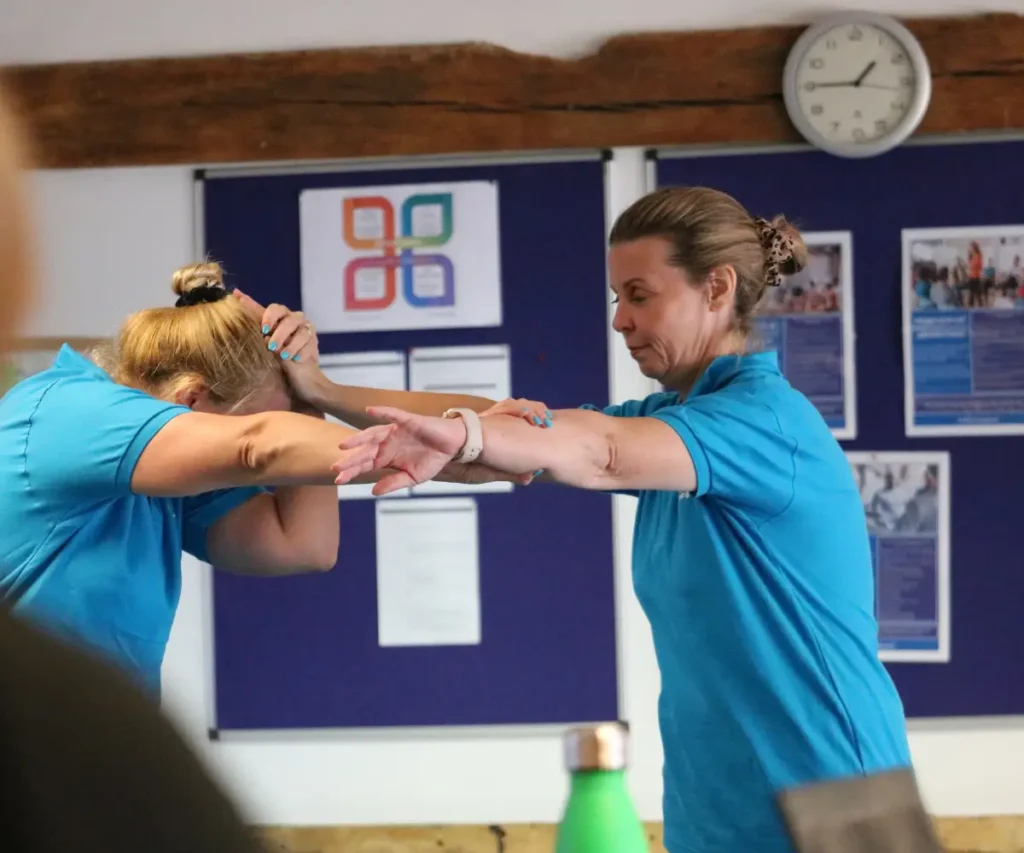 Two female instructors in blue Loddon Training polo shirts demonstrate a physical support intervention. One instructor bends forwards while the other safely guides and supports her arms and head.
