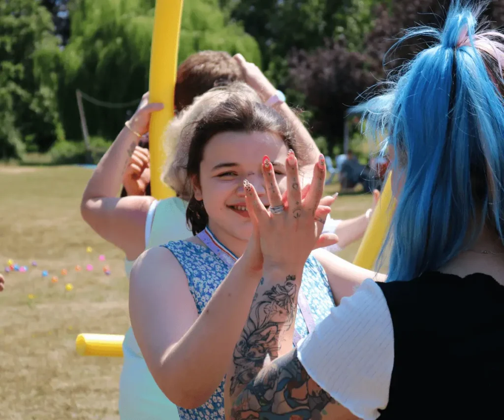 A smiling child in a blue floral dress at The Loddon School high-fives an individual with bright blue hair. They are standing outdoors in a sunny, grassy field. The person with blue hair, seen from behind, has a large floral tattoo on their forearm and small tattoos on their fingers as they raise their hand for the high-five. In the blurred background, others are participating in an activity using yellow pool noodles, with colourful small balls scattered across the grass.